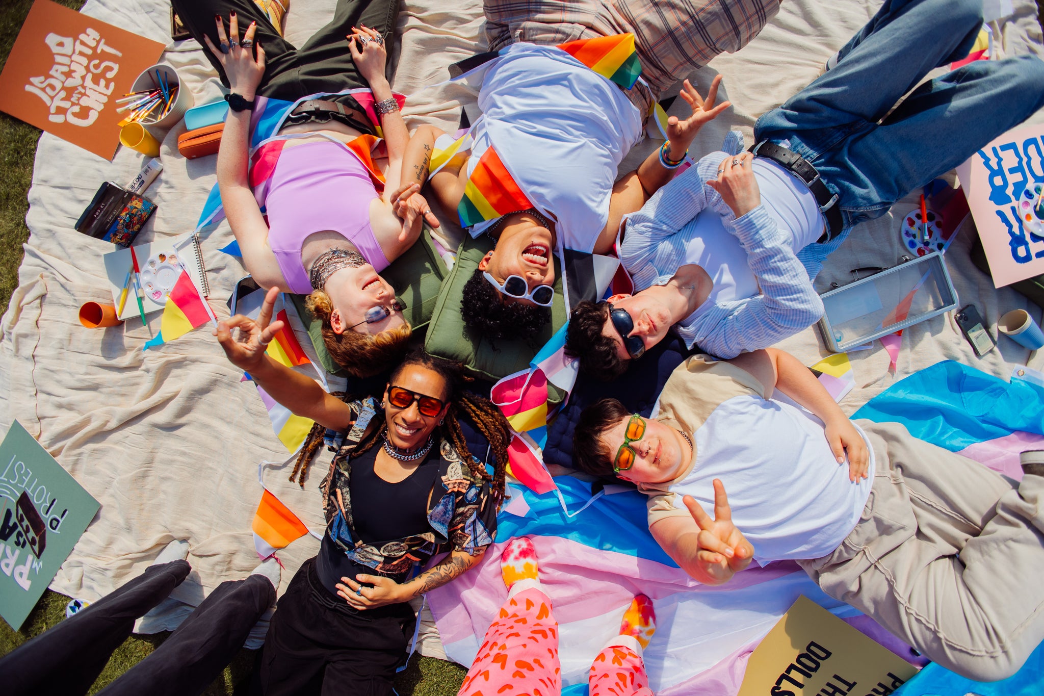 Group of lgbtqia+ friends lying on a blanket outdoors, holding hands and smiling.