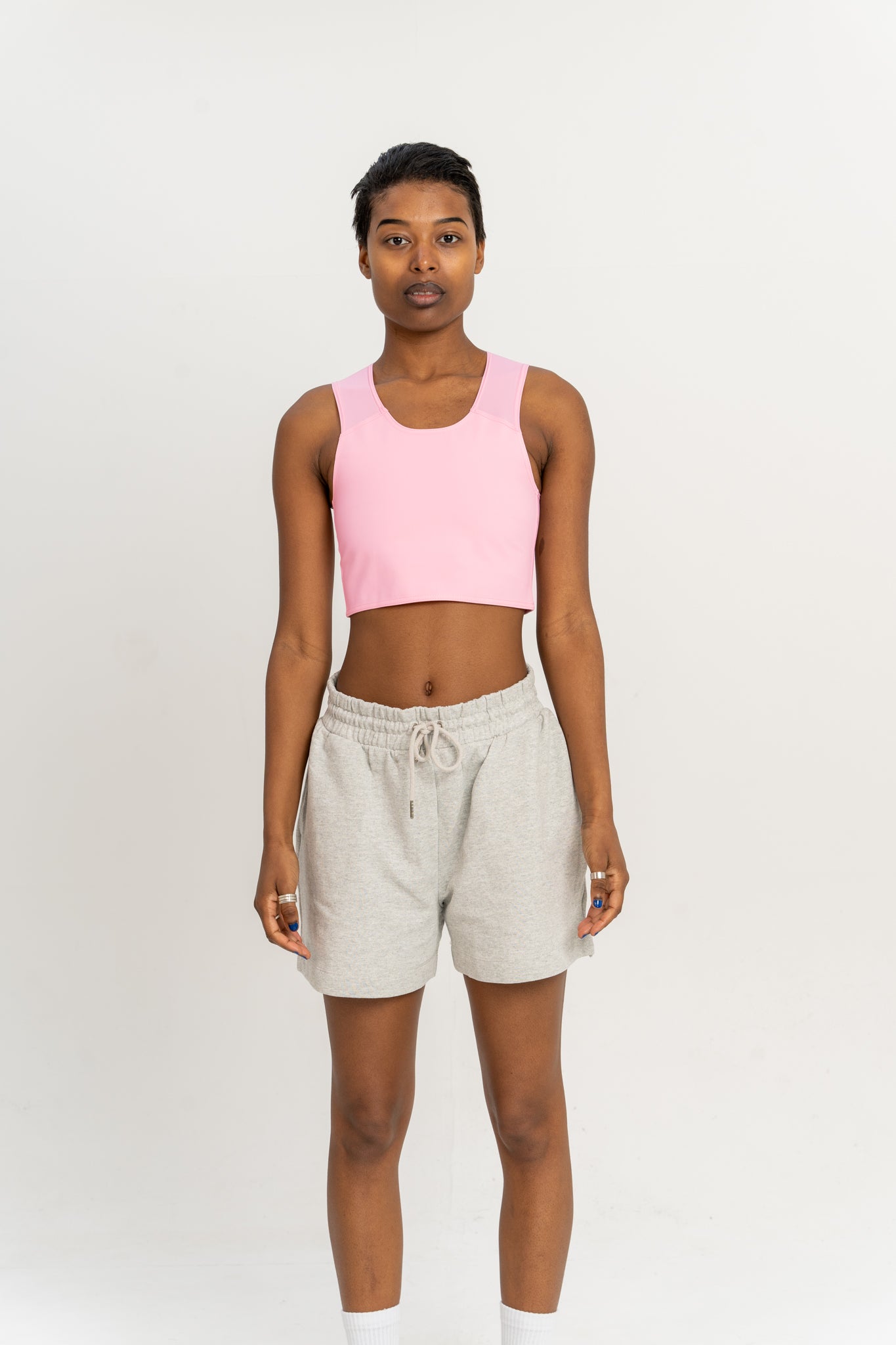 Person wearing a pastel pink coloured short chest binder and grey shorts against a white background