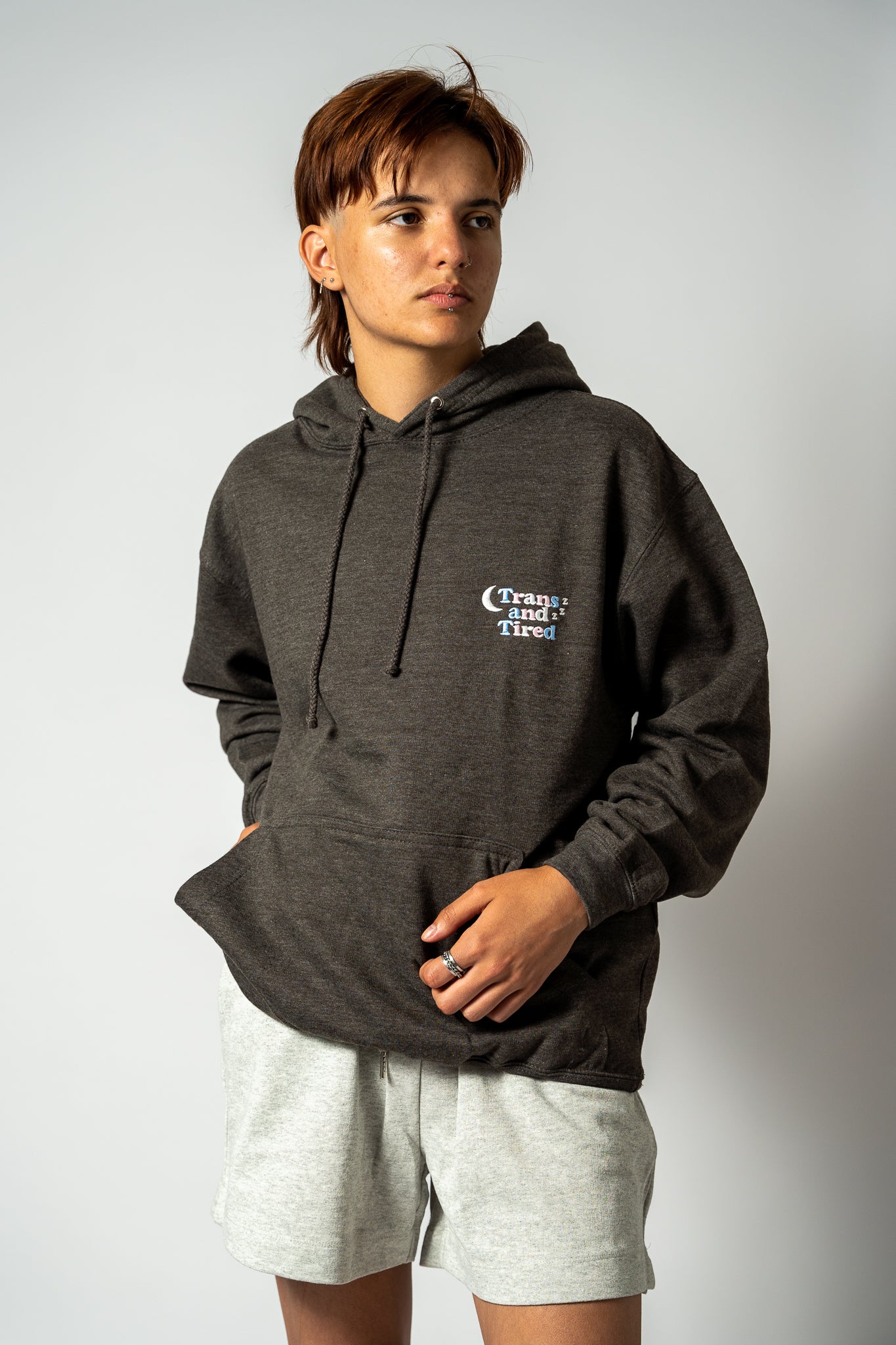Person wearing a dark gray hoodie with a 'Trans and tired' embroidery on a plain background