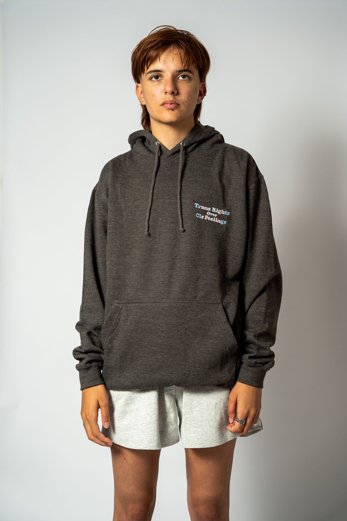 Person wearing a dark gray hoodie with 'Trans Rights over Cis Feelings' embroidery on a plain background