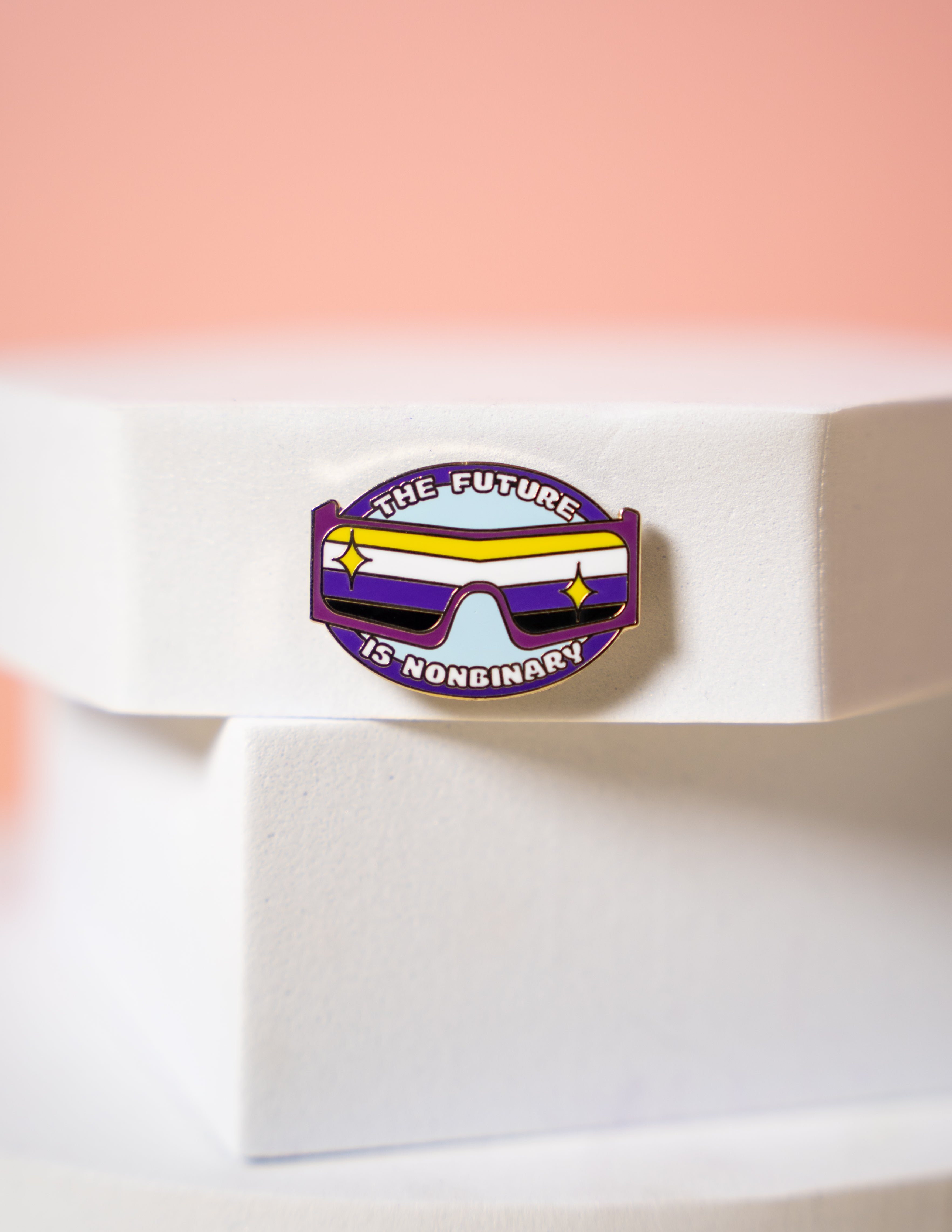 A small pin featuring non binary flag sunglasses and text reading "The future is Nonbinary"
