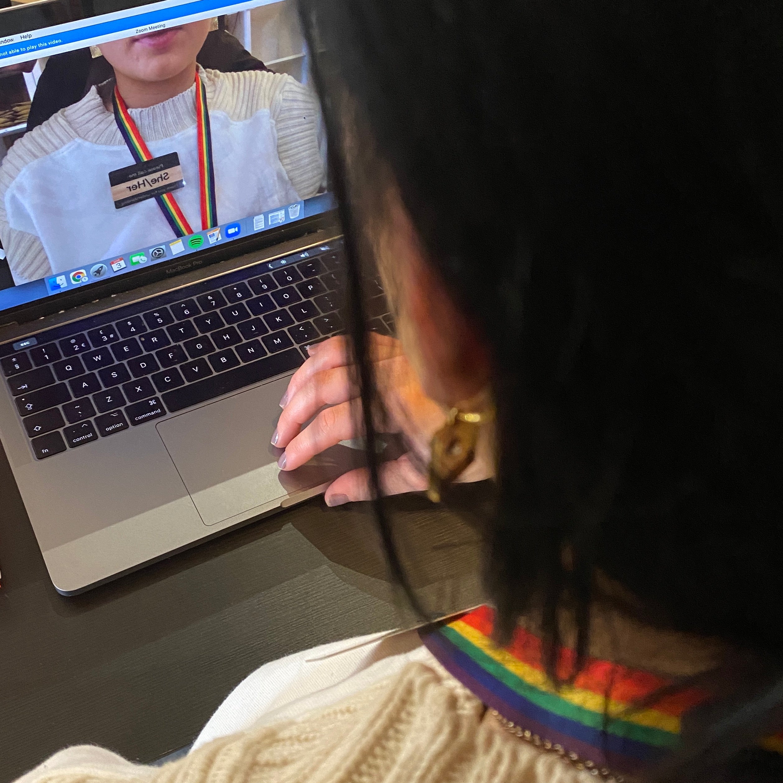 Person using a laptop with a rainbow-themed lanyard and a wooden card showing their pronouns on display.
