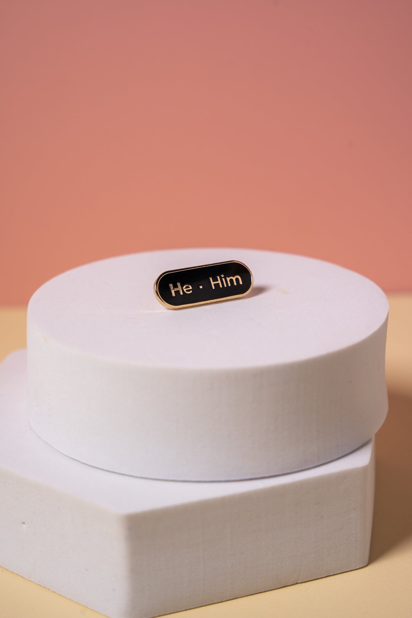 A gold and black enamel  'He Him' pronoun pin on a peach background