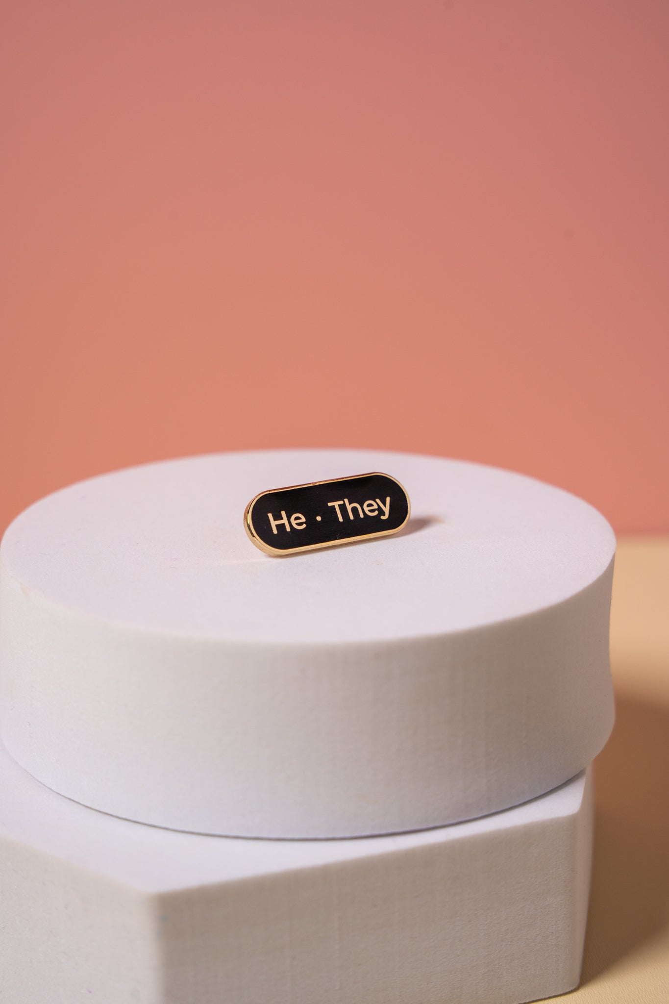A gold and black enamel  'He They' pronoun pin on a peach background