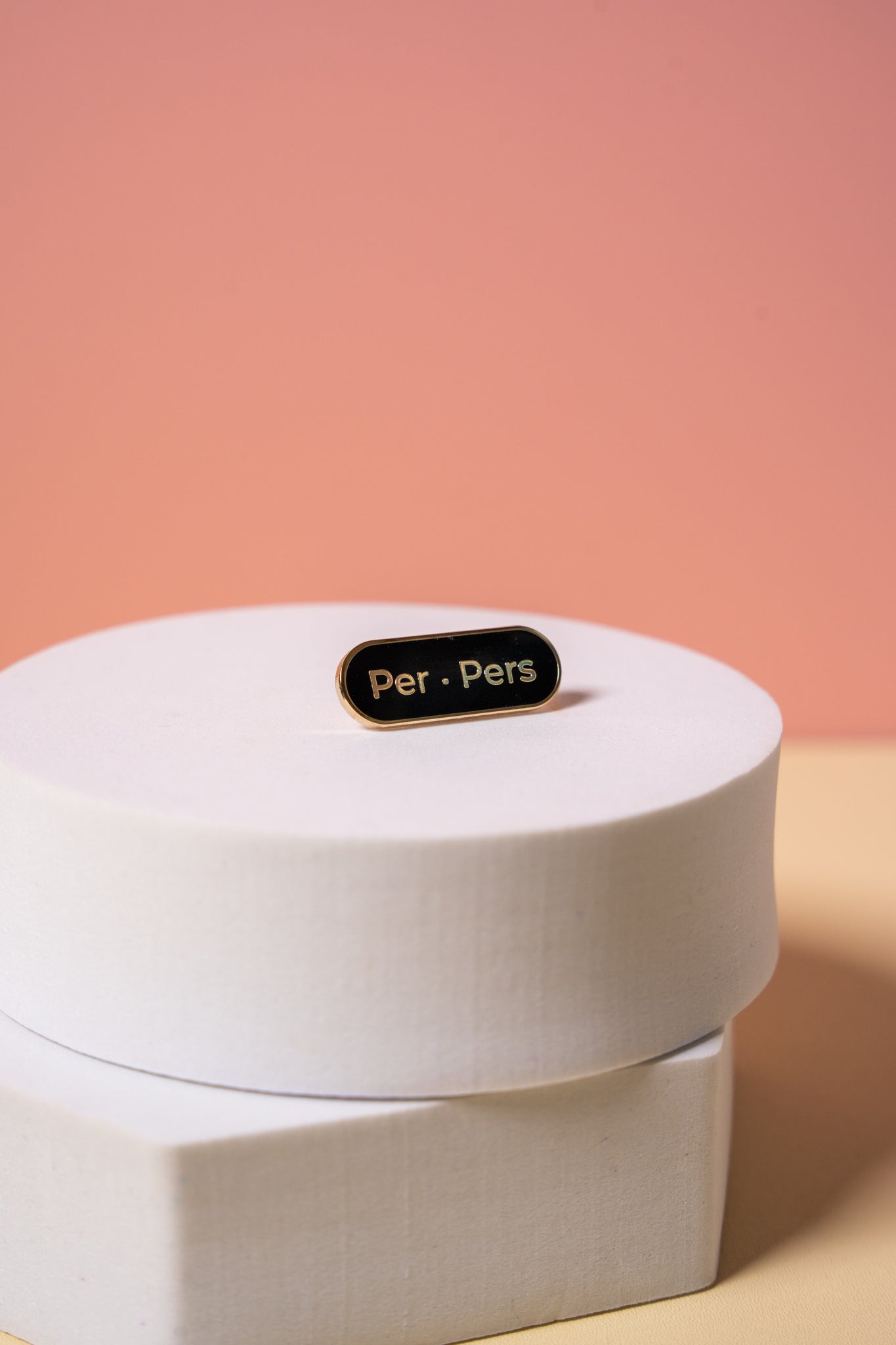 A gold and black enamel  'Per Pers' pronoun pin on a peach background
