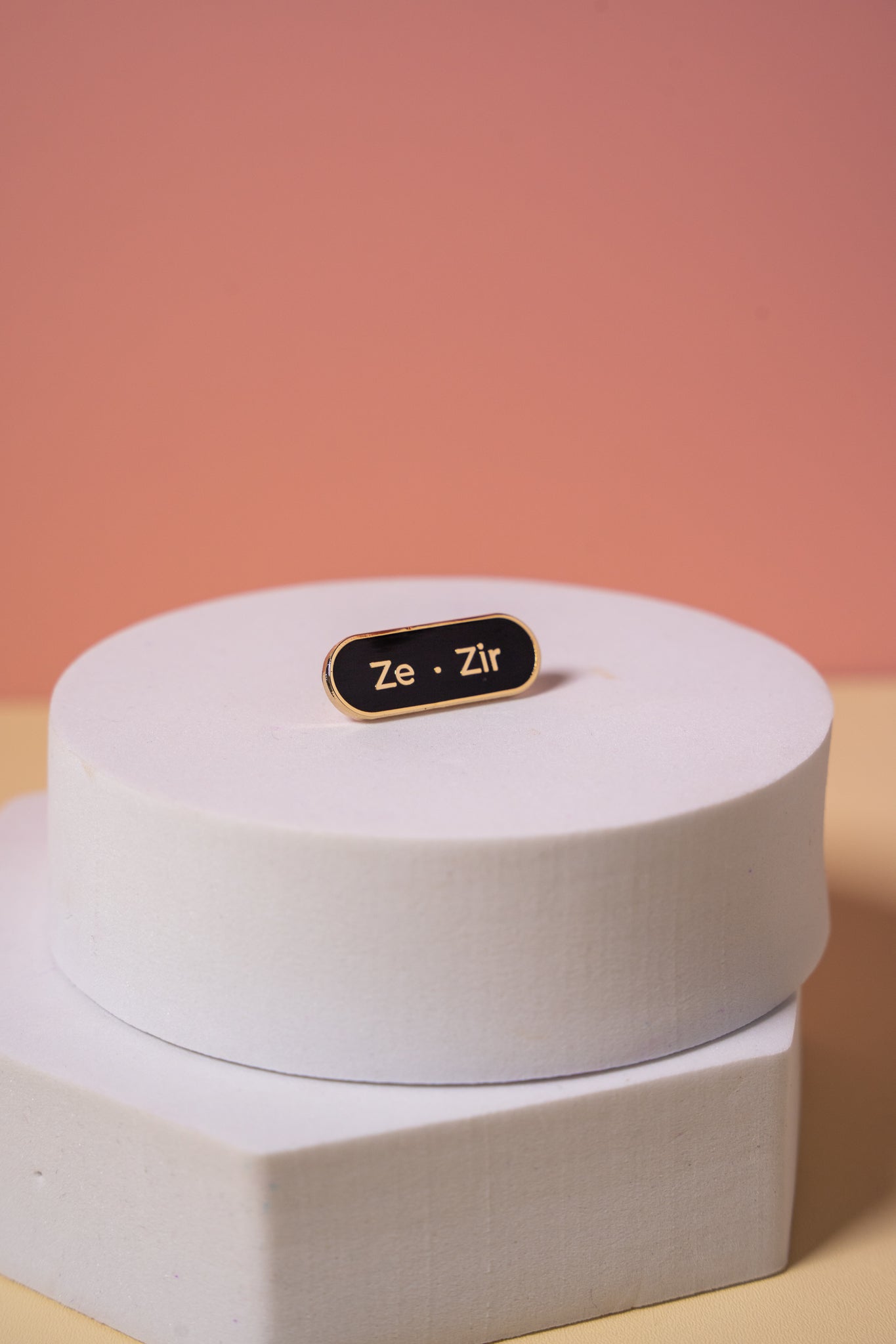 A gold and black enamel  'Ze Zir' pronoun pin on a peach background