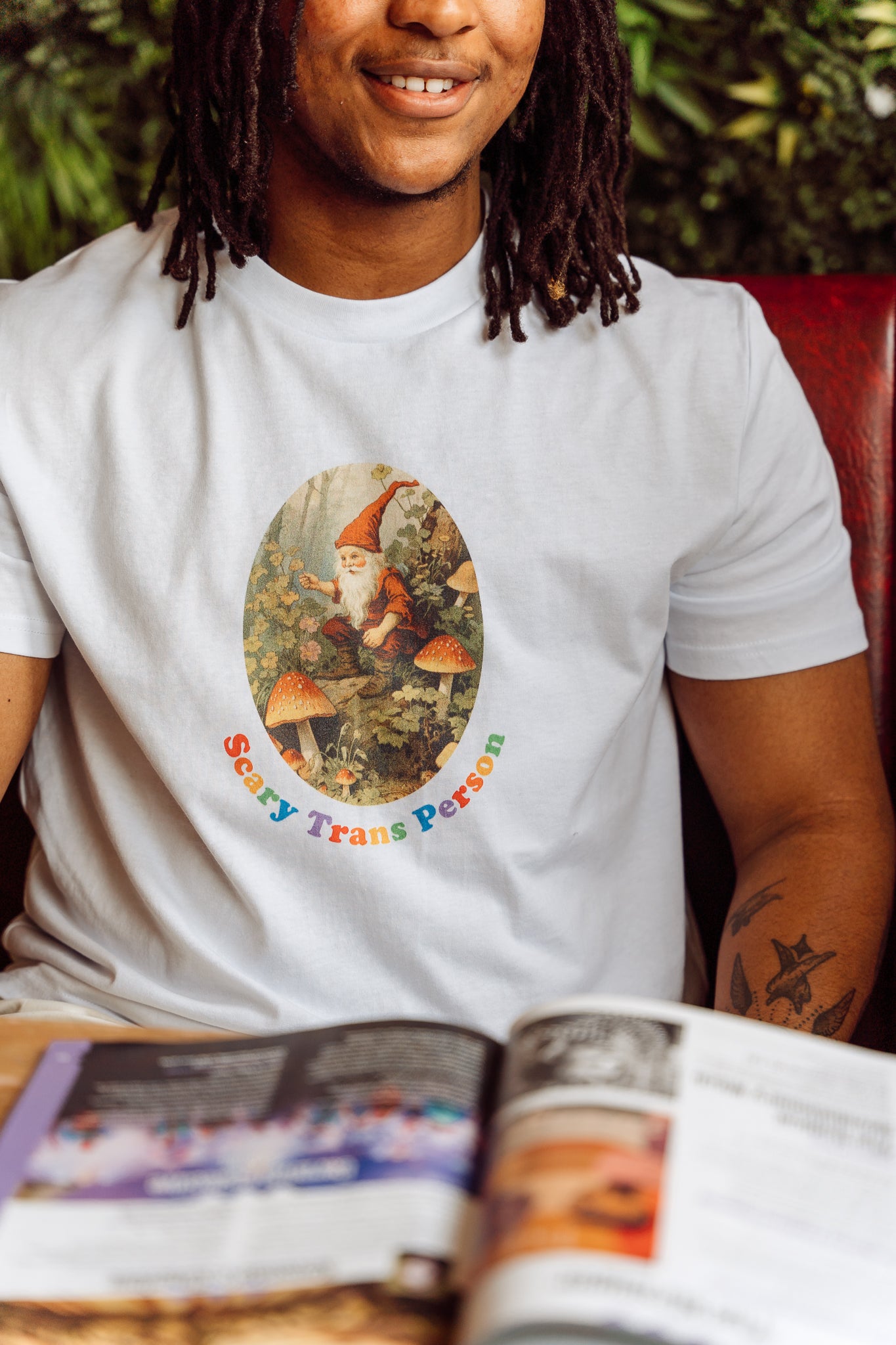 Person wearing a white t-shirt with a woodland gnome graphic featuring the text "Scary Trans Person", sitting at a table with a magazine.