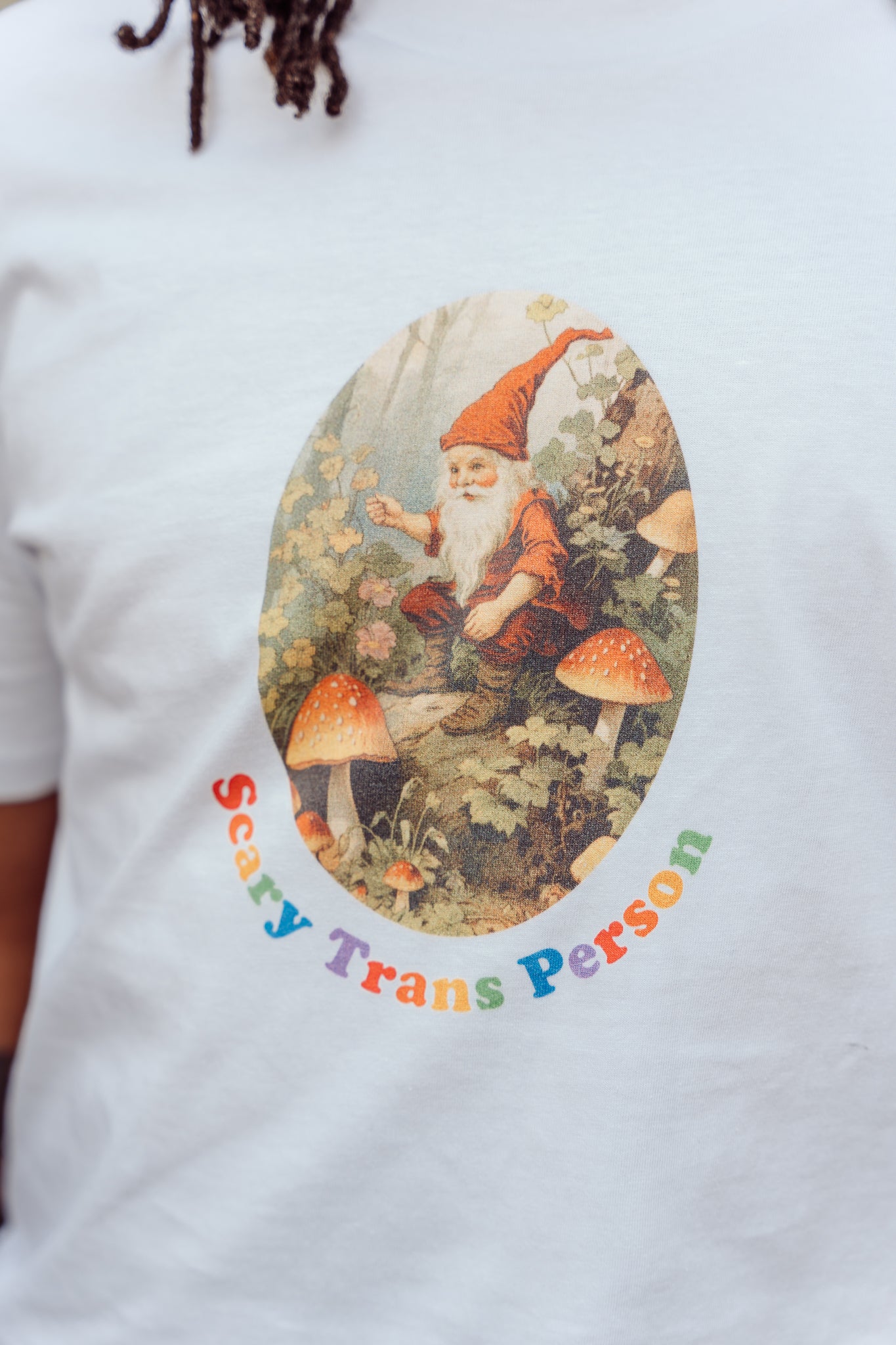 White t-shirt with a graphic of a gnome and mushrooms and text 'Scary Trans Person' on a white background.