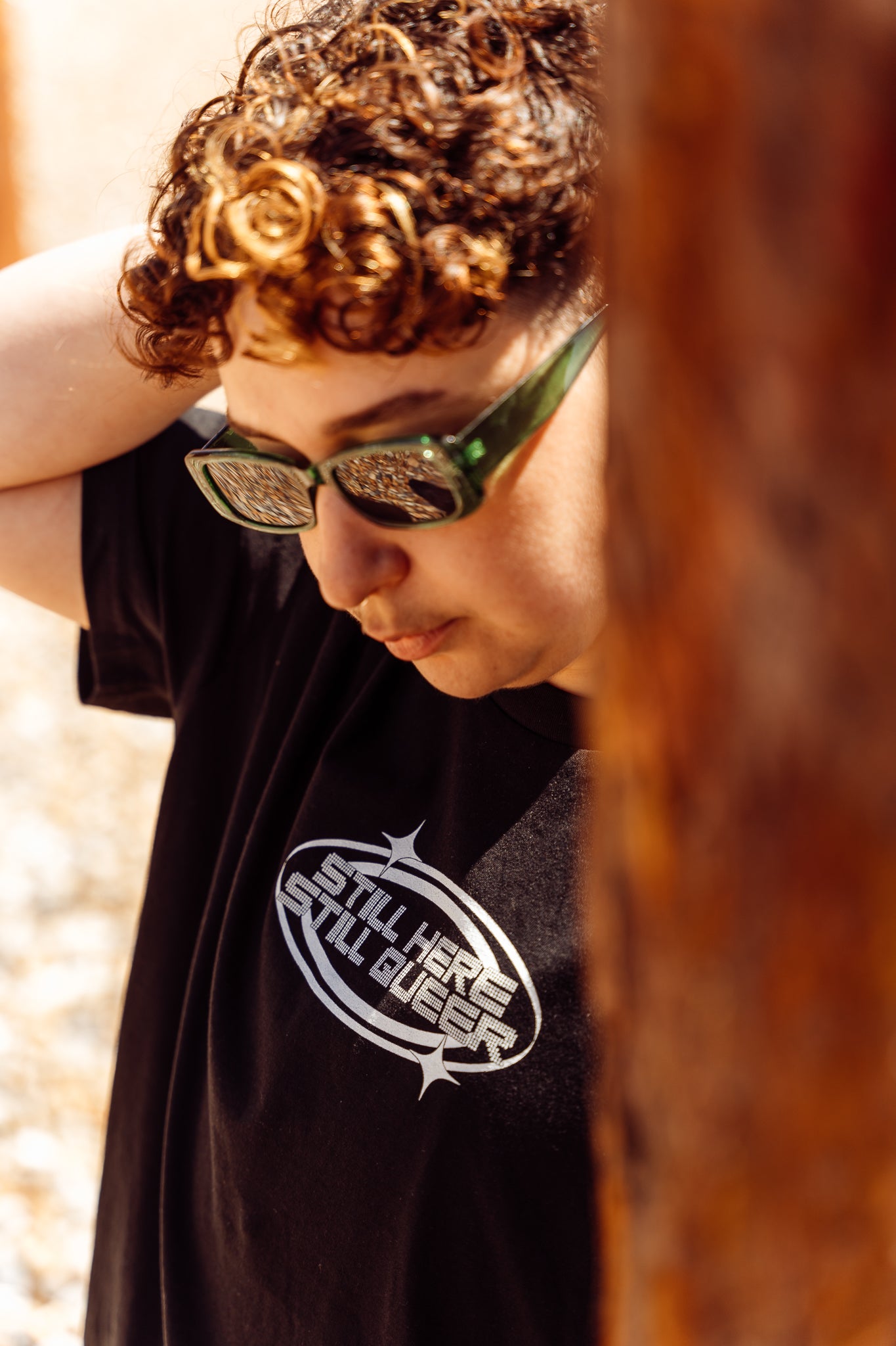 Person wearing sunglasses and a black 'Still here still queer' t-shirt, standing against a blurred natural background.